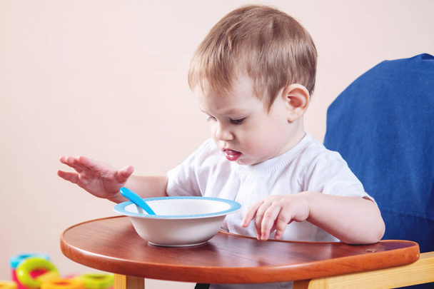 Маленький милый мальчик учится есть за детским столом, изучает тарелку и ложку на кухне.
 - Фото, изображение