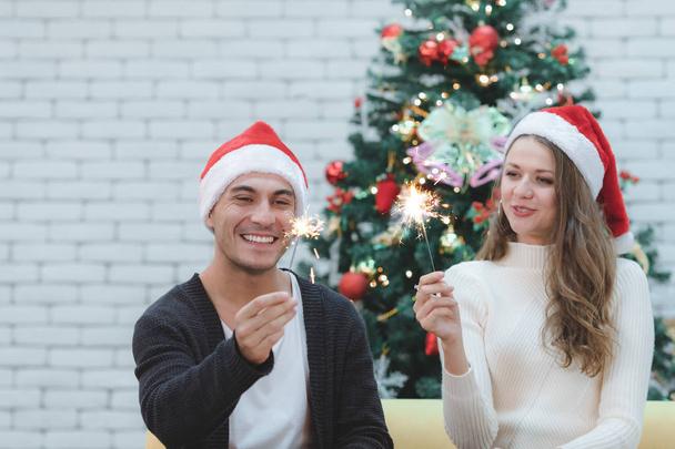 Der Fokus des jungen Paares mit rotem Hut, das auf einem Sofa zwischen Weihnachtsbäumen sitzt und gemeinsam das Mini-Feuerwerk zündet, verschwimmt. Konzept für Glück und gute gemeinsame Zeit beim Weihnachts- und Neujahrsfest. - Foto, Bild