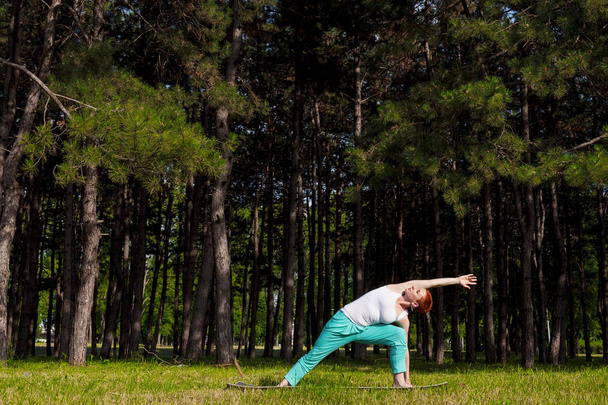 Yoga instructor outdoors-Utthita Trikonasana, Redhead girl doing Yoga in the park, trees behind, sunlit, horizontal - Photo, Image