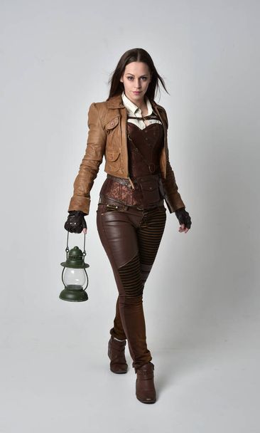 full length portrait of brunette  girl wearing brown leather steampunk outfit. standing pose holding a gas lantern on grey studio background. - Foto, Imagen