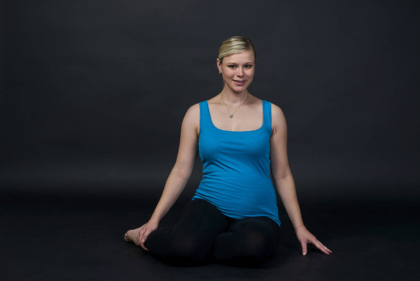 Молодая блондинка беременная женщина в синей майке делает упражнения, черный фон
 - Фото, изображение