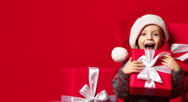 Mosolygós vicces gyerek Mikulás piros kalapban, kezében karácsonyi ajándékkal. Karácsonyi koncepció. - Fotó, kép