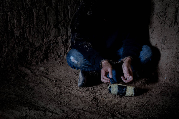 Traficante de drogas bajo arresto confinado con esposas, sentado junto a una pared. Enfócate en el paquete de drogas
 - Foto, Imagen