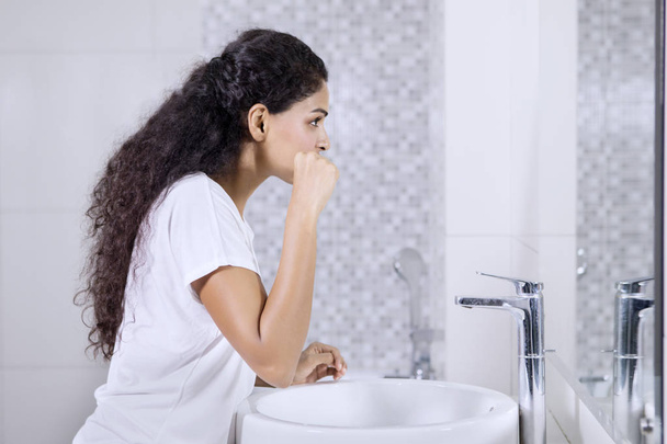 Side view of Indian woman brushing teeth while standing in the bathroom - Fotó, kép