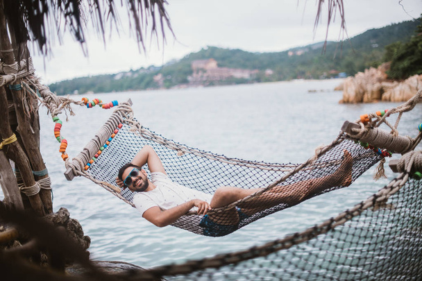 Молодий чоловік лежить у гамаку біля океану. Красивий хлопець відпочиває в гамаку на тлі гір біля моря
. - Фото, зображення