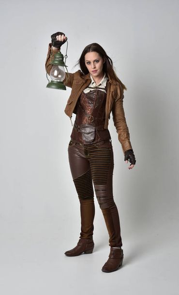 Portret van de volledige lengte van brunette meisje bruinleren steampunk outfit dragen. staande pose houden een gas lantaarn op grijs studio achtergrond. - Foto, afbeelding