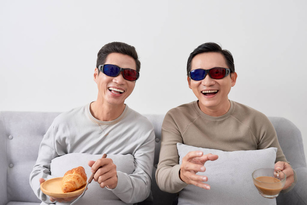 Два азиата в 3D очках сидят на диване и смотрят кино
 - Фото, изображение