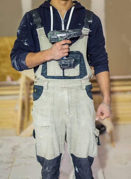 Worker in dirty uniform or boilersuit  with drill in his hand in the room of apartment  that  is under construction, remodeling, renovation, extension, overhaul, restoration and reconstruction  - Фото, зображення