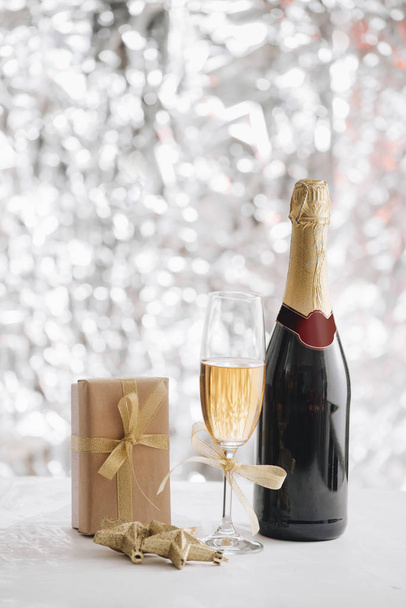 бокал шампанского и бутылка с подарочной коробкой и рождественские украшения на серебряном фоне блесток
 - Фото, изображение