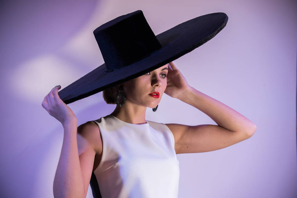 Mujer atractiva joven de pie sobre el fondo de una pared blanca en sombrero grande. Iluminación morada
 - Foto, imagen