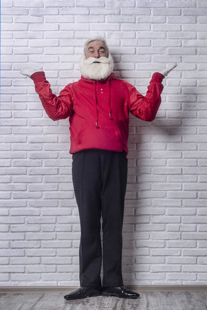 A grey-haired bearded man in a red jacket in the image of Santa Claus is standing against a white wall and posing emotionally. - 写真・画像