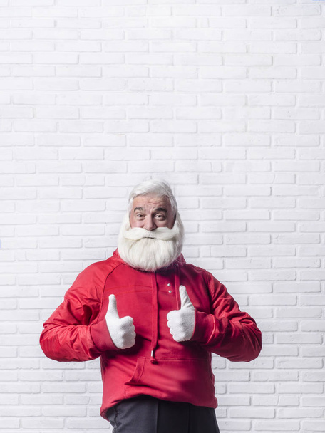 A grey-haired bearded man in a red jacket in the image of Santa Claus is standing against a white wall and posing emotionally. - Foto, Imagem