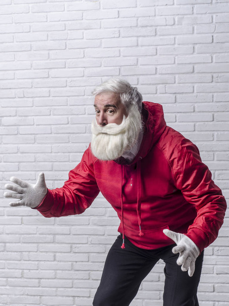 A grey-haired bearded man in a red jacket in the image of Santa Claus is standing against a white wall and posing emotionally. - Foto, Bild
