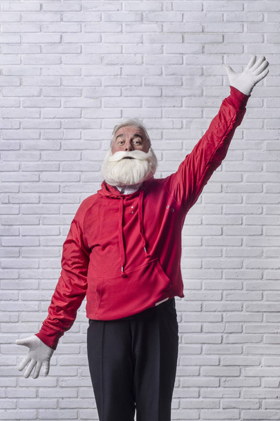 A grey-haired bearded man in a red jacket in the image of Santa Claus is standing against a white wall and posing emotionally. - Foto, Bild