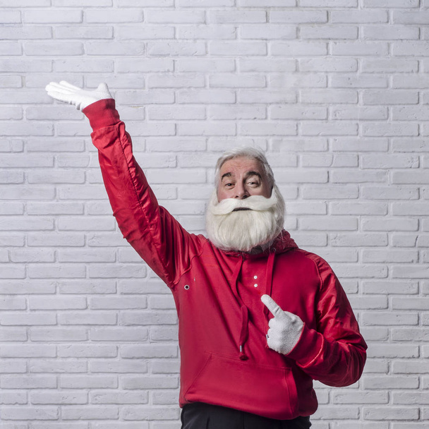 A grey-haired bearded man in a red jacket in the image of Santa Claus is standing against a white wall and posing emotionally. - Фото, зображення
