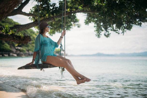 Een jonge vrouw is swingend op een schommel in de schaduw van een grote boom door de zee. Meisje op de schommel strand aan de kust van een tropisch eiland. - Foto, afbeelding
