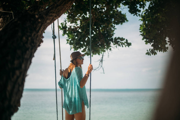 A young woman is swinging on a swing in the shade of a big tree by the sea. Girl on the beach swing on the coast of a tropical island. - Φωτογραφία, εικόνα