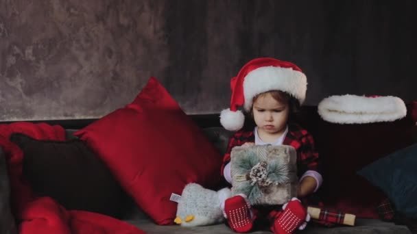 Child holding and opening a Christmas gift box, wearing in Santa hat. Kid having fun at home. Xmas winter holiday concept - Séquence, vidéo