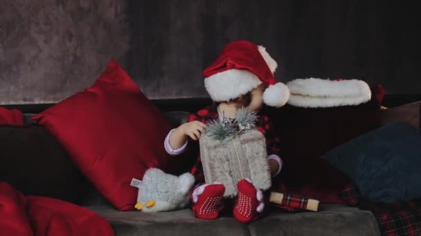 Child holding and opening a Christmas gift box, wearing in Santa hat. Kid having fun at home. Xmas winter holiday concept - Materiał filmowy, wideo