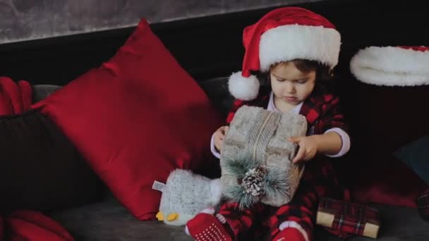 Child holding and opening a Christmas gift box, wearing in Santa hat. Kid having fun at home. Xmas winter holiday concept - Metraje, vídeo