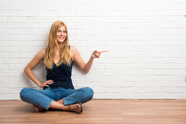 Blonde girl sitting on the floor pointing finger to the side and presenting a product on white brick wall background - Photo, Image