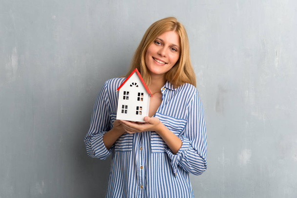 Блондинка тримає маленький будинок на текстурованому гранжевому фоні стіни
 - Фото, зображення