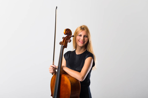 Молодая блондинка со своей виолончелью позирует на белой стене
 - Фото, изображение