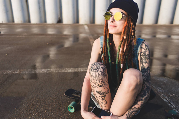 young girl with tattoos and dreadlocks in a blue cap sits on a longboard against the background of concrete structure - Foto, afbeelding