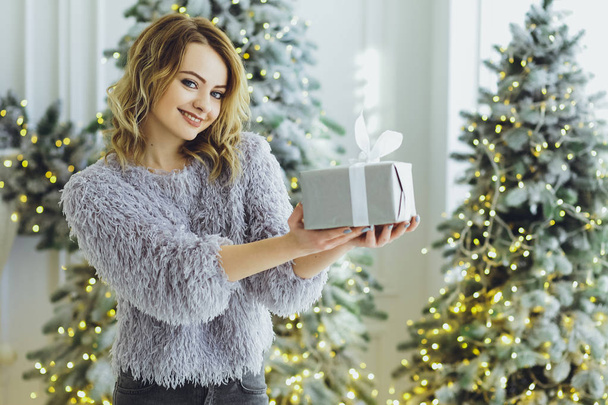 Young beautiful blonde girl in a gray sweater with a gift on hands near the New Year, Christmas tree with gifts - Foto, immagini