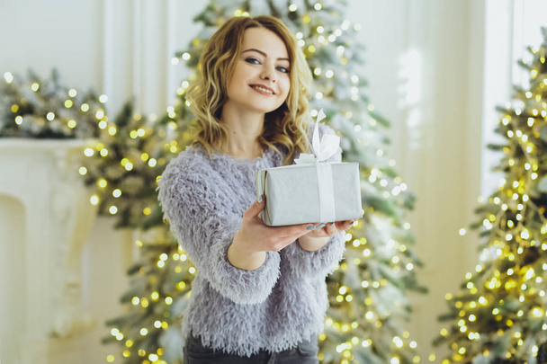 Young beautiful blonde girl in a gray sweater with a gift on hands near the New Year, Christmas tree with gifts - Фото, зображення
