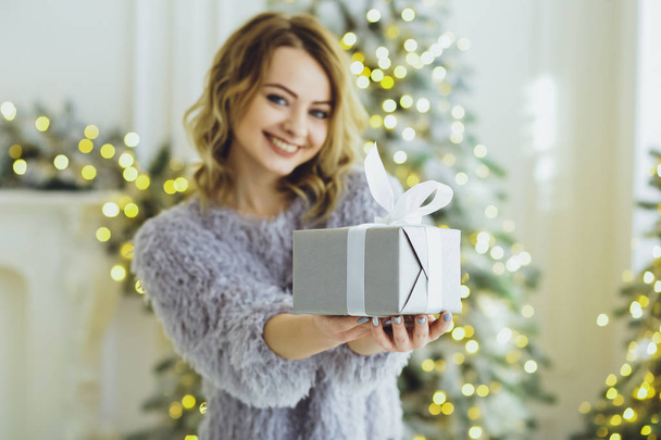 Young beautiful blonde girl in a gray sweater with a gift on hands near the New Year, Christmas tree with gifts - Foto, imagen