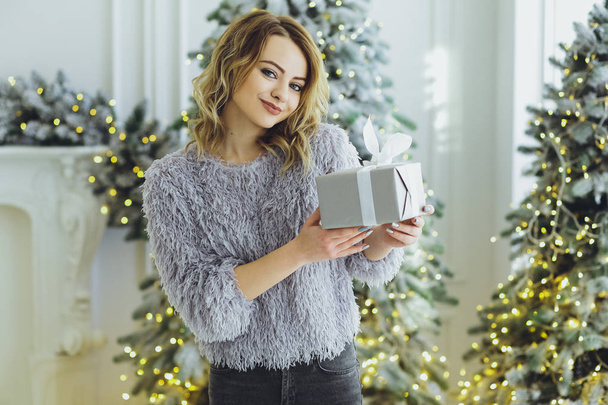 Young beautiful blonde girl in a gray sweater with a gift on hands near the New Year, Christmas tree with gifts - Fotoğraf, Görsel