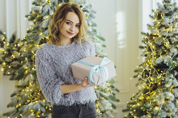 Young beautiful blonde girl in a gray sweater with a gift on hands near the New Year, Christmas tree with gifts - Foto, Imagen