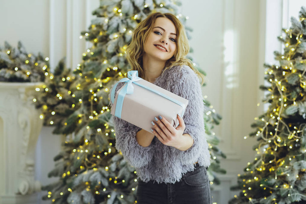 Young beautiful blonde girl in a gray sweater with a gift on hands near the New Year, Christmas tree with gifts - Foto, Imagem