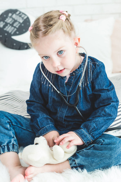 Cute little girl sitting on the bed and playing doctor with stethoscope and Teddy Bear. - Foto, Bild