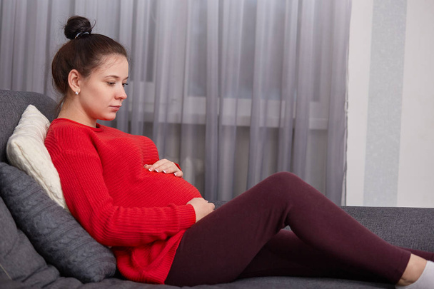 Horizontal shot of relaxed future mother wears red knitted sweater, leggings, sits at couch, has thoughtful expression, keeps hands on tummy, awaits for baby. Pregnancy and domestic atmosphere - Zdjęcie, obraz