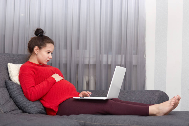 Photo of expectant woman keeps hand on belly, being pregnant, spends leisure time at home, watches exciting movie on portable laptop compuer, poses at sofa, wears comfortable domestic clothes - Foto, afbeelding