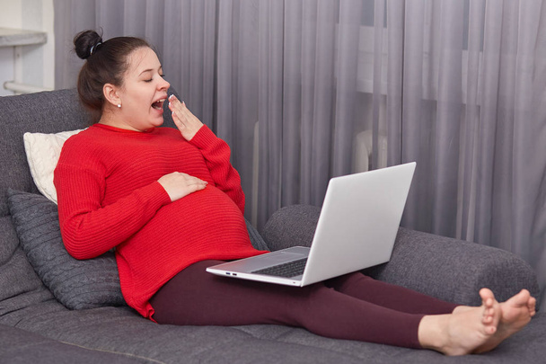 Tired expecting mother yawns as wants to sleep, works freelance at laptop computer, dressed in casual domestic clothes, sits at sofa, covers mouth with palm. Pregancy, weariness, home concept - 写真・画像