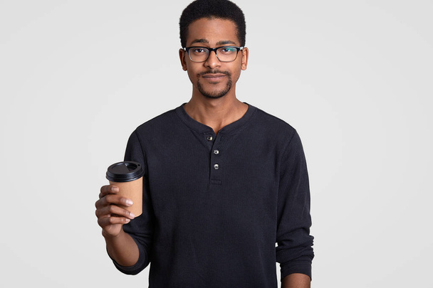 Horizontal shot of serious black young man holds takeaway coffee, wears transparent glasses, has break after work, models against white background, enjoys spare time. People and drinking concept - Фото, зображення
