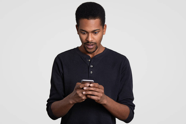 Astonished black man with curly hair, holds mobile phone, recieves text message, feels surprised and amazed, wears casual clothes, isolated over white background. People, reaction and technology - Valokuva, kuva