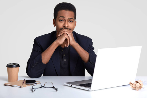 Photo of sleepy tired prosperous male business owner keeps hands under chin, needs to develop new strategy to raise profit, dressed formally, uses laptop computer and cellular, isolated on white wall - 写真・画像