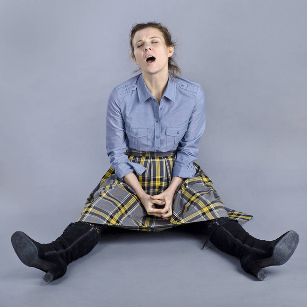 A beautiful European woman is funny and originally sits on the floor. A beautiful European woman is funny and originally sits on the floor in a blue shirt and a checkered skirt on a blue background. - Fotoğraf, Görsel