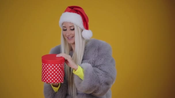 Happy beautiful smiling woman in fur coat and Santa hat opening a red gift box on yellow background. Christmas celebrate, wow effect and surprise - Záběry, video