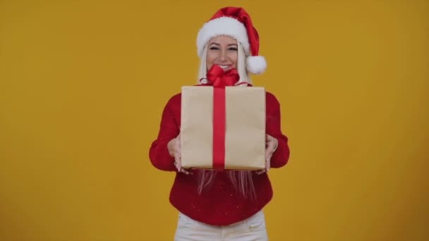 Felice eccitato giovane ragazza in cappello di Babbo Natale Riceve un regalo - una scatola con un nastro rosso brillante su sfondo giallo isolato
.  - Filmati, video