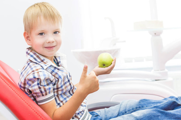 Dental clinic. Reception, examination of the patient. Teeth care. Little boy holding an apple while sitting in a dental chair - Photo, image