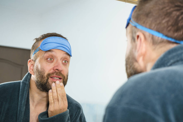 Funny adult man wearing eyemask on forhead having troubles with waking up. Standing in front of mirror looking at his face, yawning, being tired and sleepy. - Foto, afbeelding