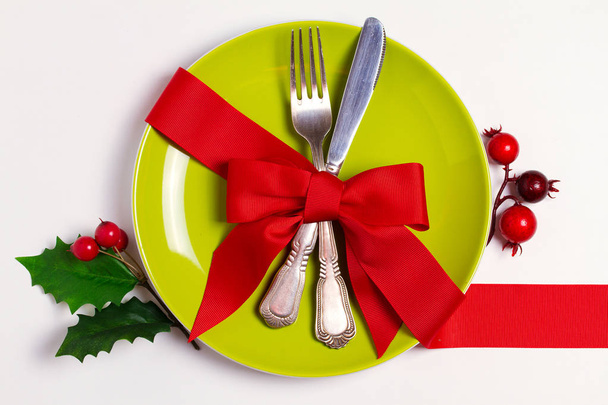 Christmas composition with plate, cutlery, pine branches, ribbon and red berries on white table. Winter holidays and festive background. Christmas eve dinner, New Year food lunch. View from above, top - Foto, Bild