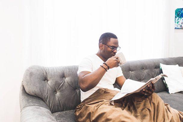 Young African American man, covered with a blanket reading a newspaper at home. - Фото, изображение