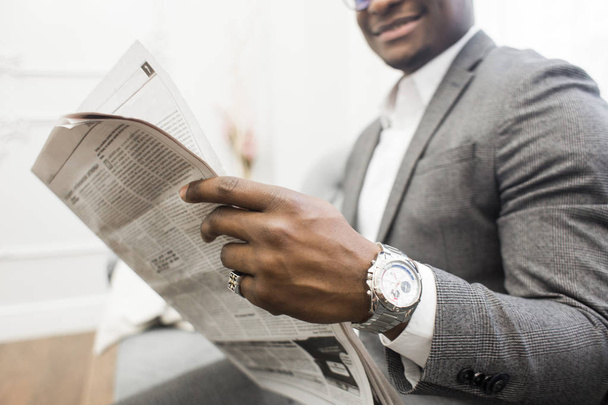 Young African American businessman in a gray suit reading a newspaper while sitting on a sofa. - Φωτογραφία, εικόνα