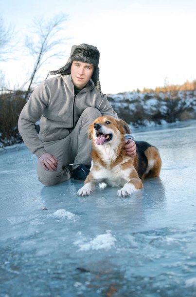 young man on a walk with a dog sitting on the first transparent winter ice - Photo, Image
